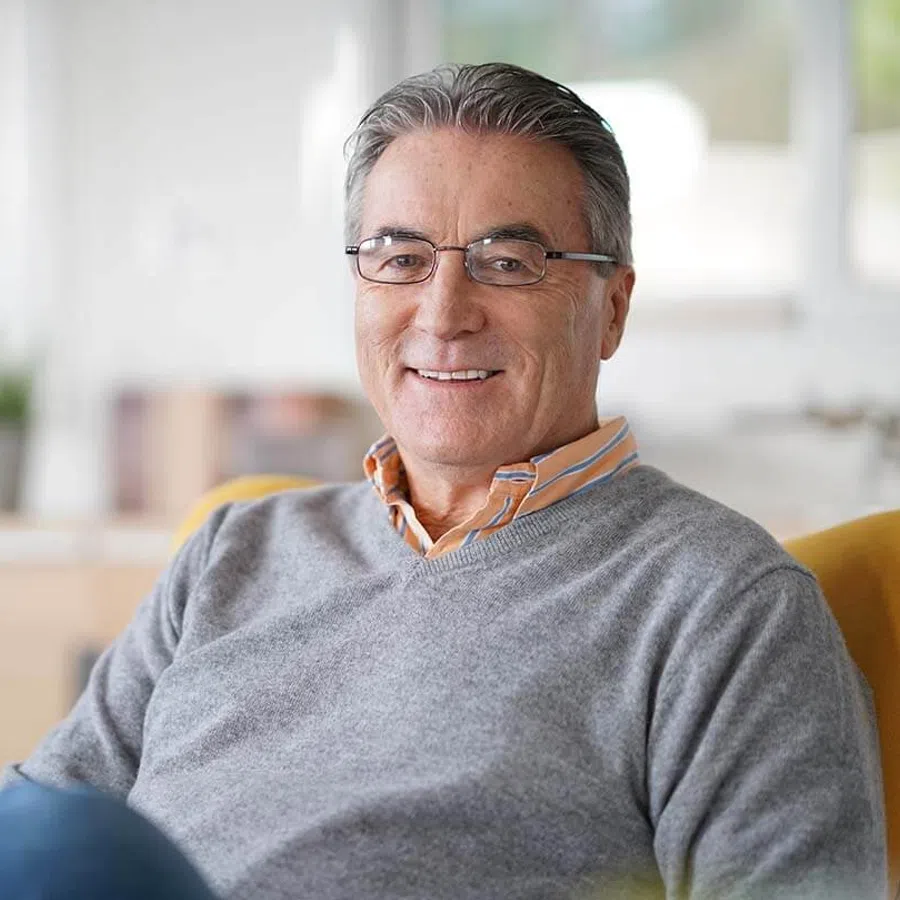 Smiling middle-aged man wearing glasses and a gray sweater sitting indoors in a bright room.