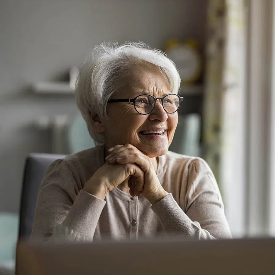 Smiling elderly woman with white hair and glasses sitting indoors, hands clasped, looking out a window.
