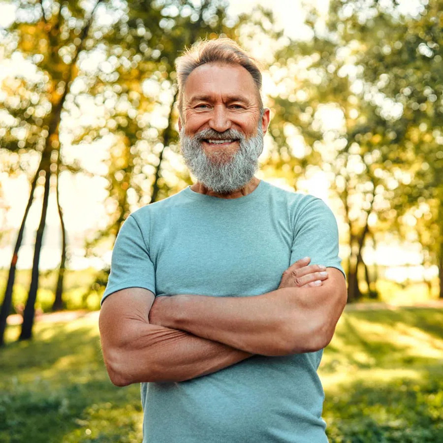 Smiling middle-aged man with gray beard and folded arms standing outdoors in a sunlit green park.