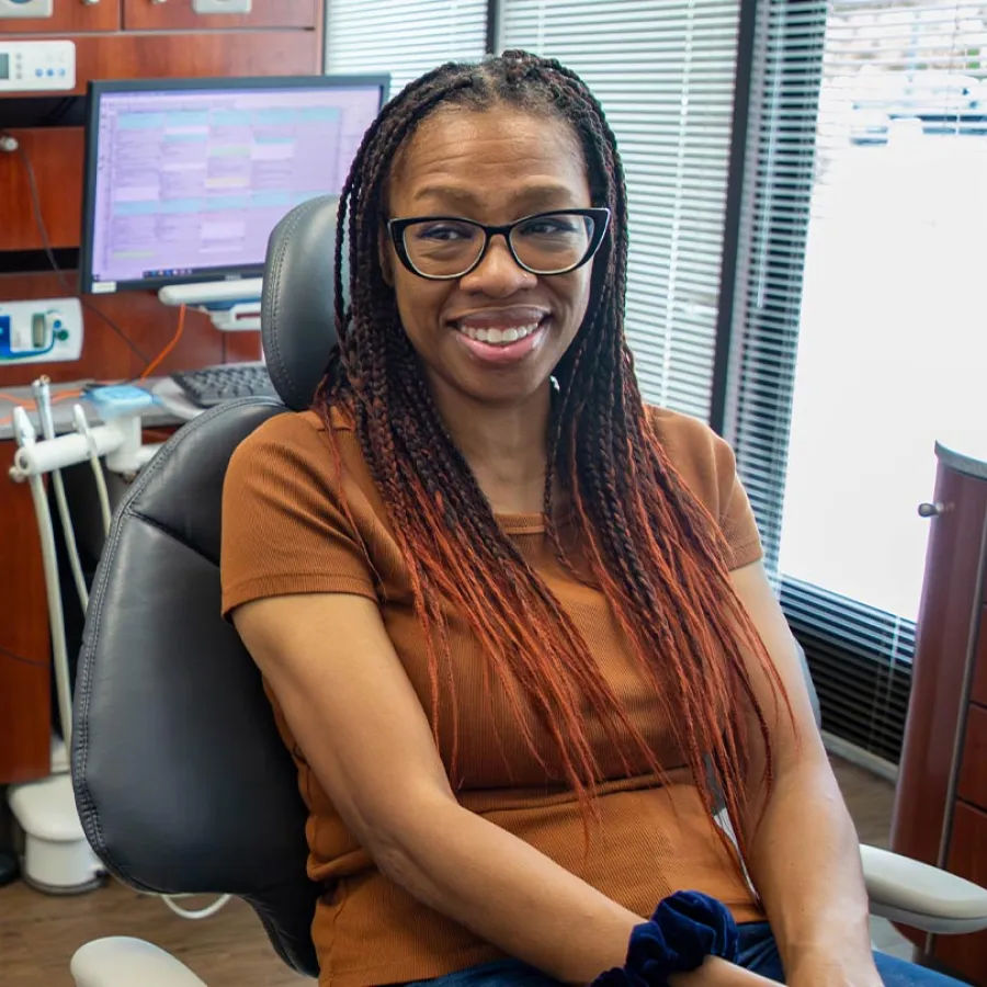 Smiling woman with glasses and braided hair sitting in a dental office chair near computer and equipment.