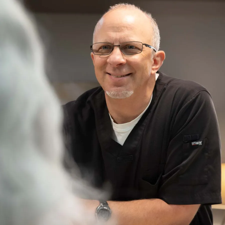 Smiling male healthcare professional wearing glasses and black scrubs talking to a patient with gray hair.