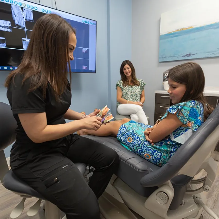 Dentist explaining dental care using a model to a young girl sitting in a dental chair with her mother watching.