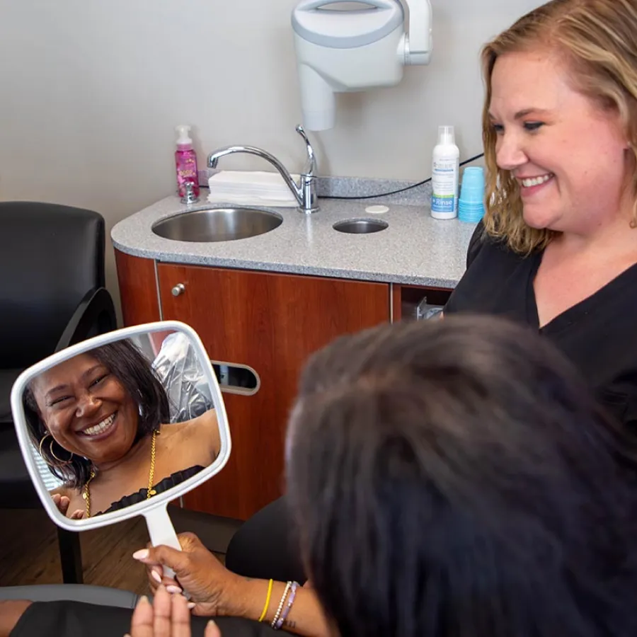 Smiling woman looks at her reflection in a hand mirror while a healthcare professional watches with a smile.