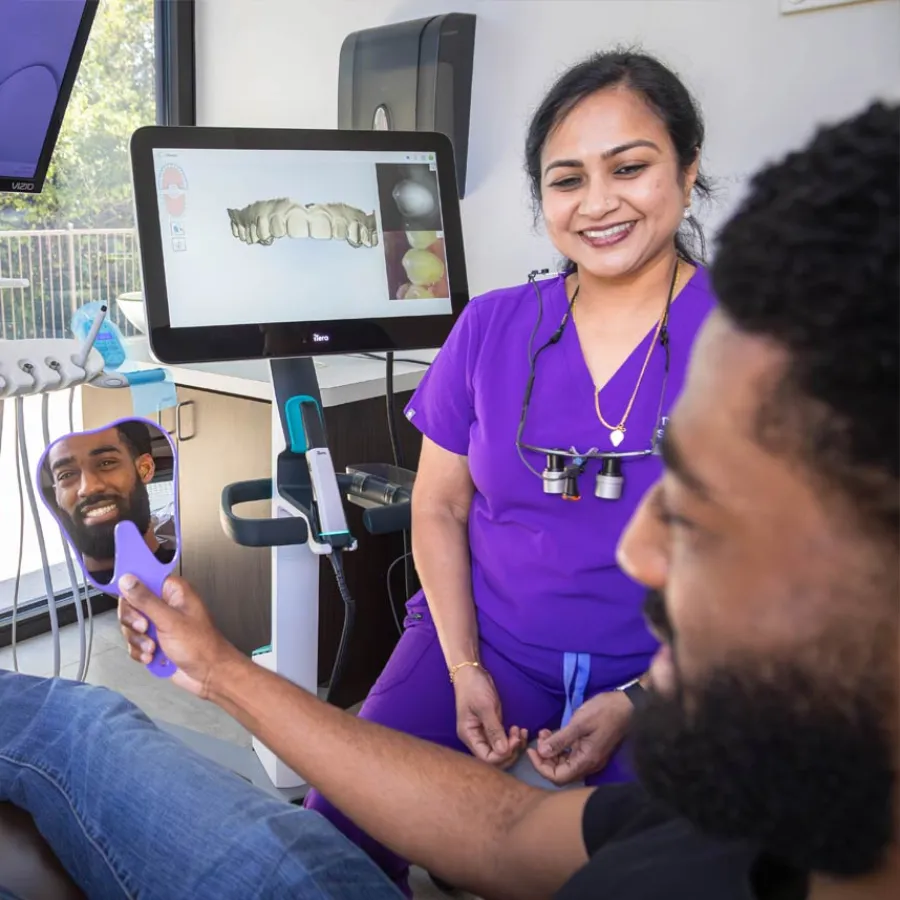 Dentist in purple scrubs smiling at patient holding mirror showing his teeth and dental 3D scan on screen