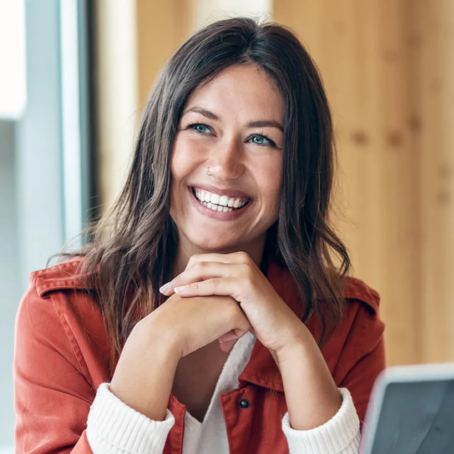 Smiling young woman with blue eyes and brown hair wearing red jacket sitting indoors by window.