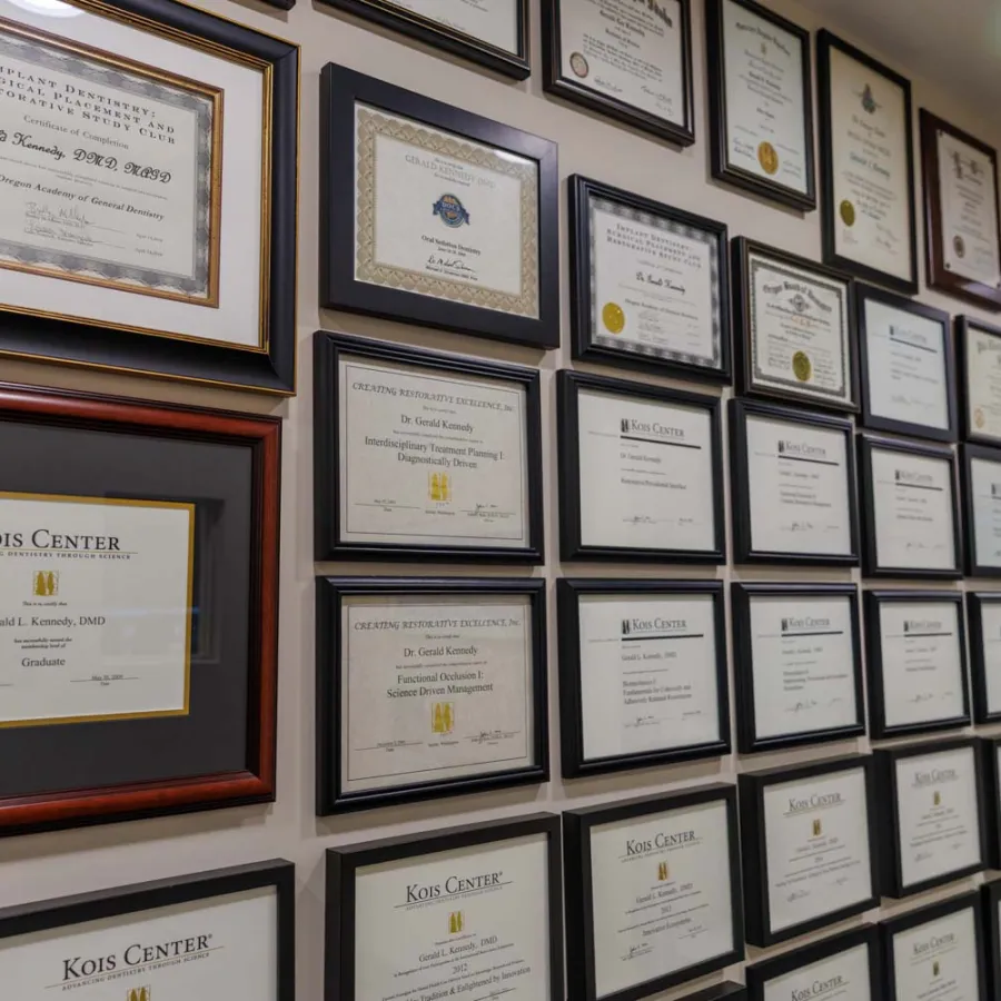 Wall displaying numerous framed certificates and diplomas in an office hallway under bright lighting.