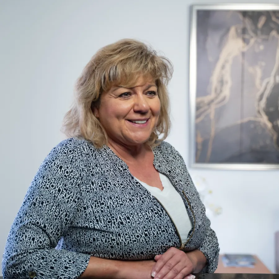 Smiling middle-aged woman in patterned jacket standing indoors with framed art in the background.