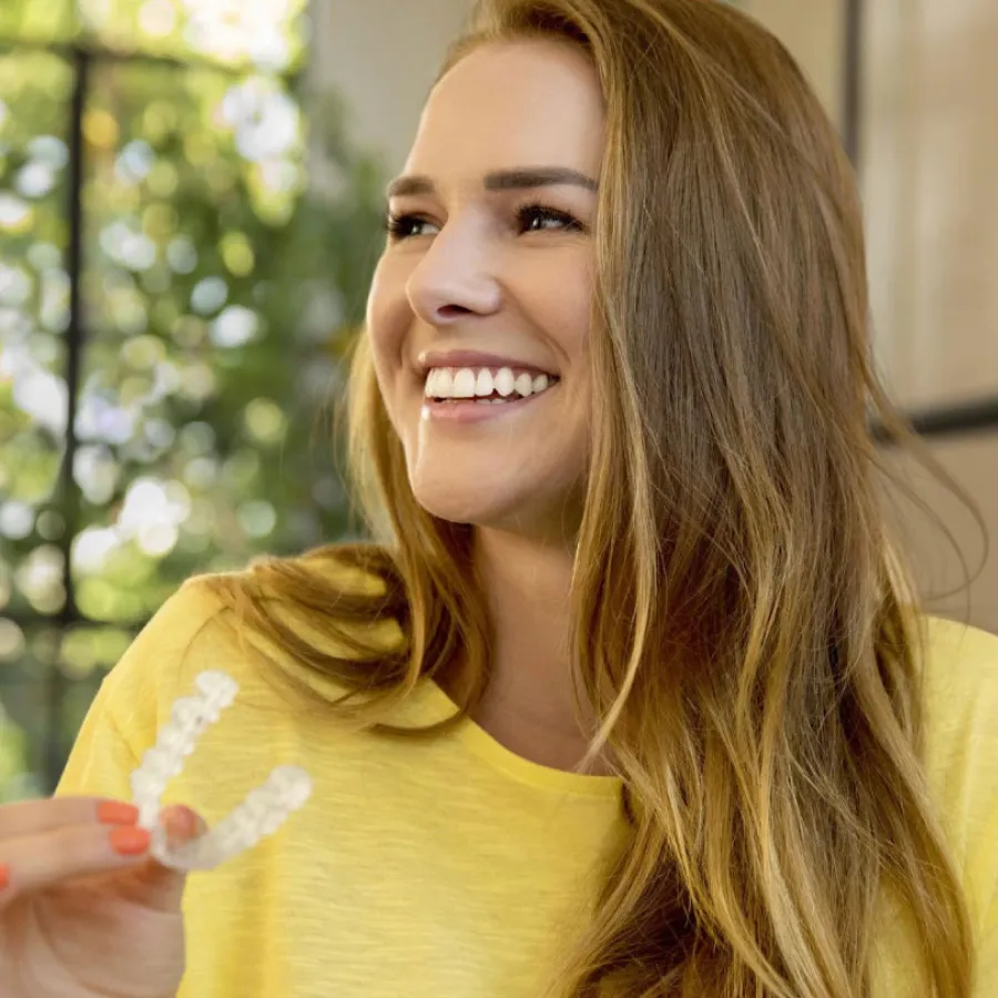 Smiling woman in yellow shirt holding a clear dental aligner with greenery blurred in the background