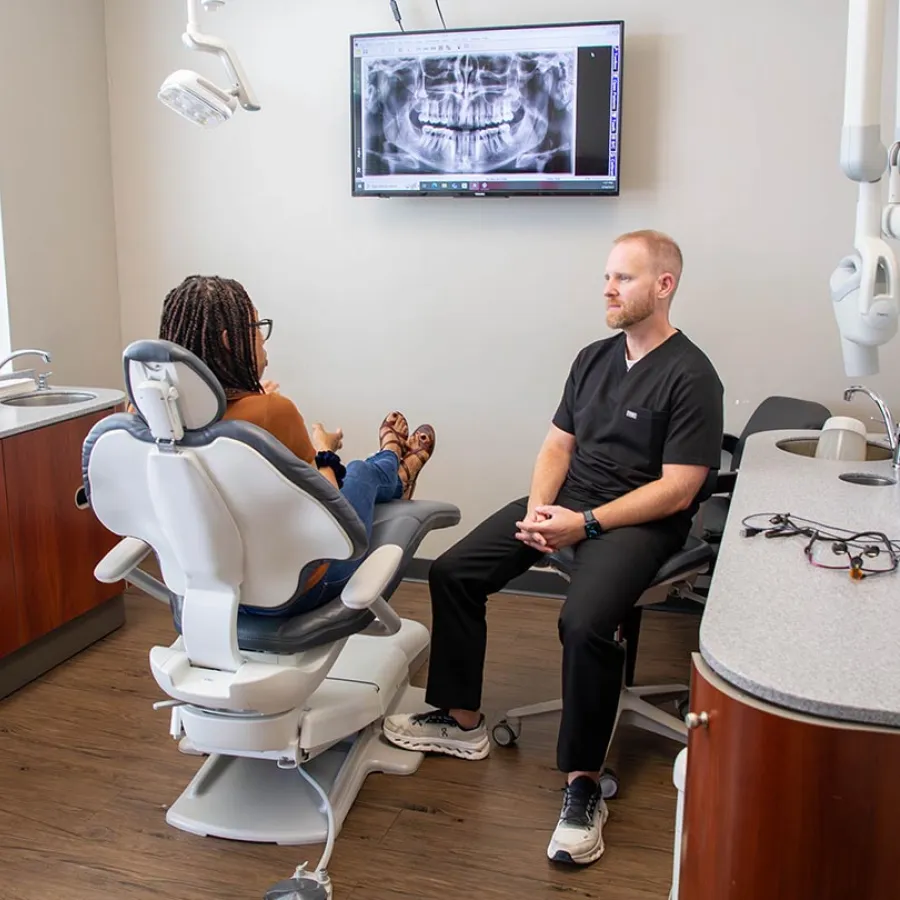Dentist consulting patient in dental office with a large digital dental X-ray image displayed on the wall.