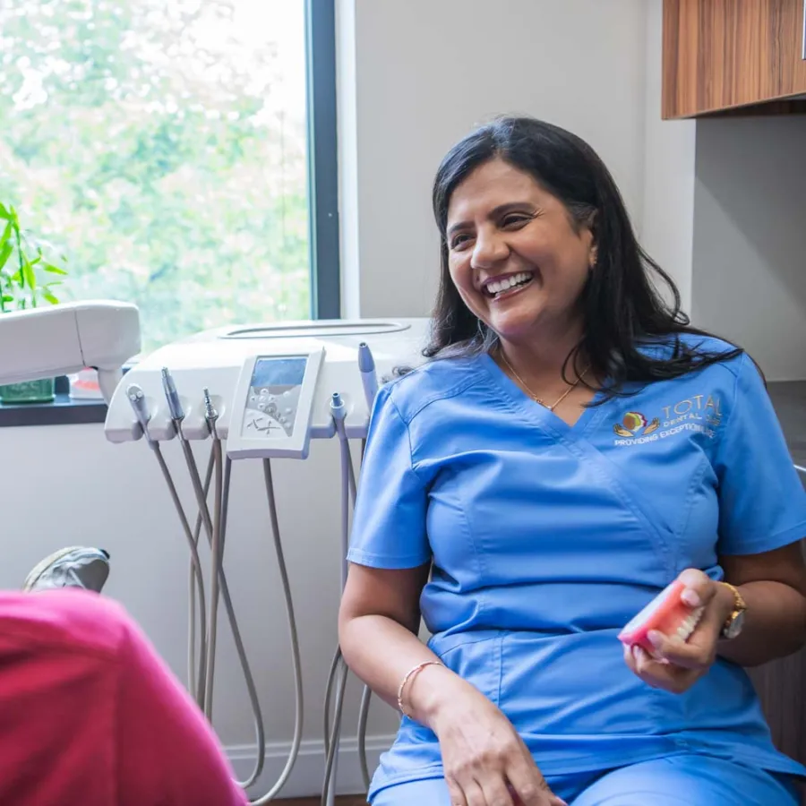 Dentist in blue scrubs smiling and talking to patient in dental office with equipment and window view.