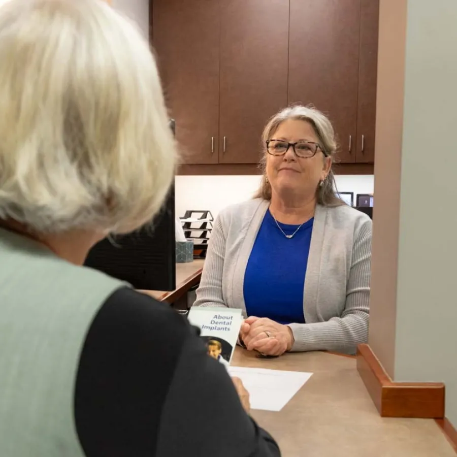 Woman in glasses and blue shirt consulting another woman about dental implants in an office setting.