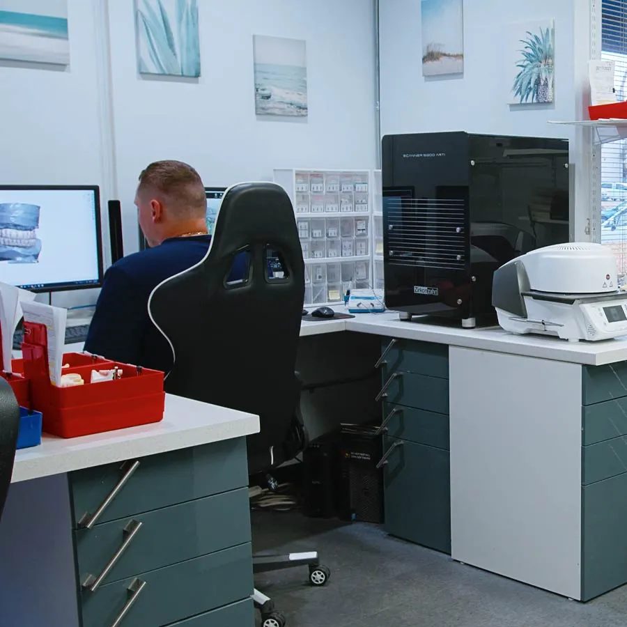 Dental technician working on 3D dental model using computer and dental equipment in clinic workspace.