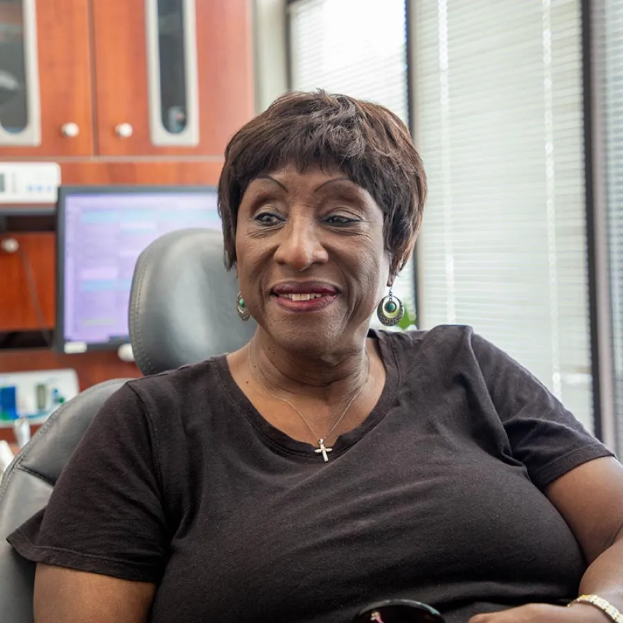 Smiling woman with short hair wearing a black shirt and cross necklace sitting in an office chair.