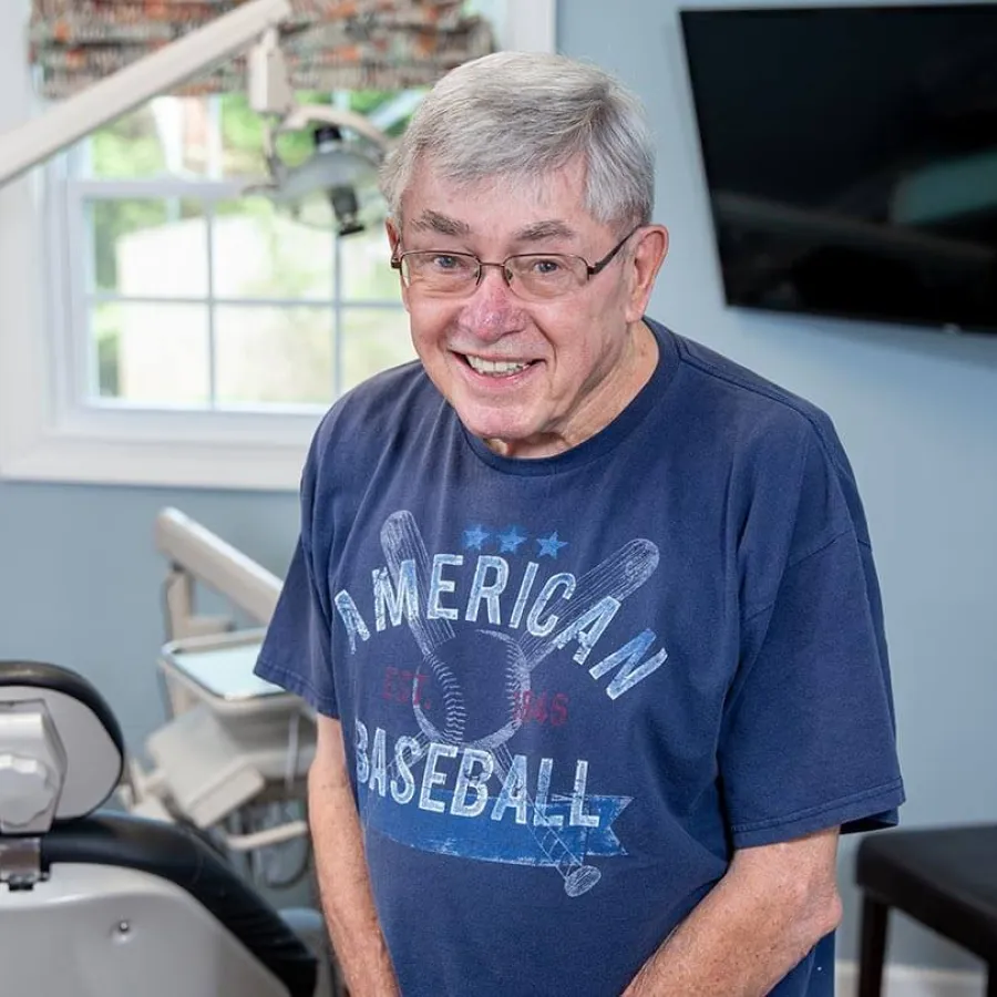 Elderly man wearing an American Baseball t-shirt smiling in a bright room with dental equipment.