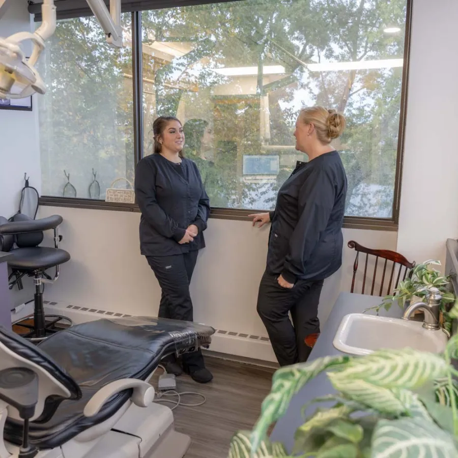 Two dental professionals in black uniforms talking in a modern dental exam room with a chair and large window.