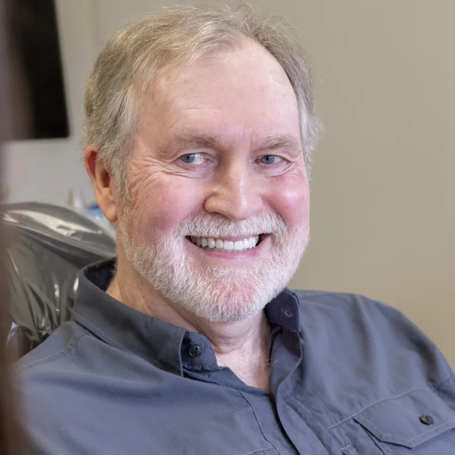 Smiling elderly man with gray beard sitting in a chair wearing a blue button-up shirt, facing a blurred person.