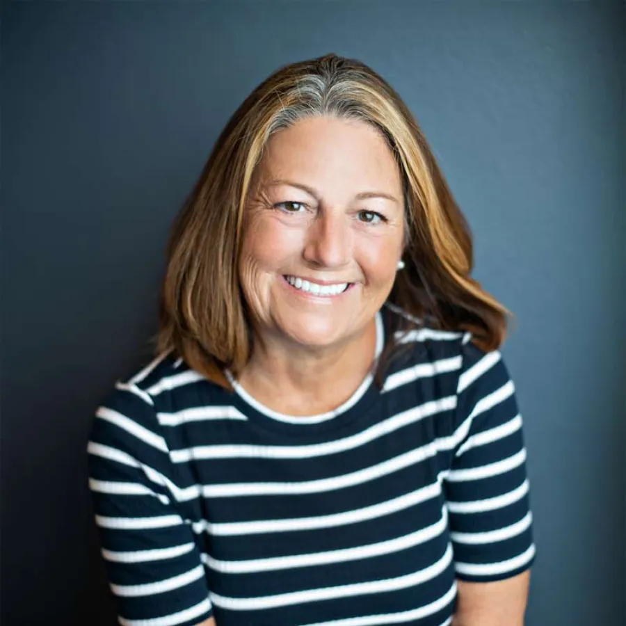 Smiling middle-aged woman with shoulder-length brown and blonde hair wearing a black and white striped shirt against dark background