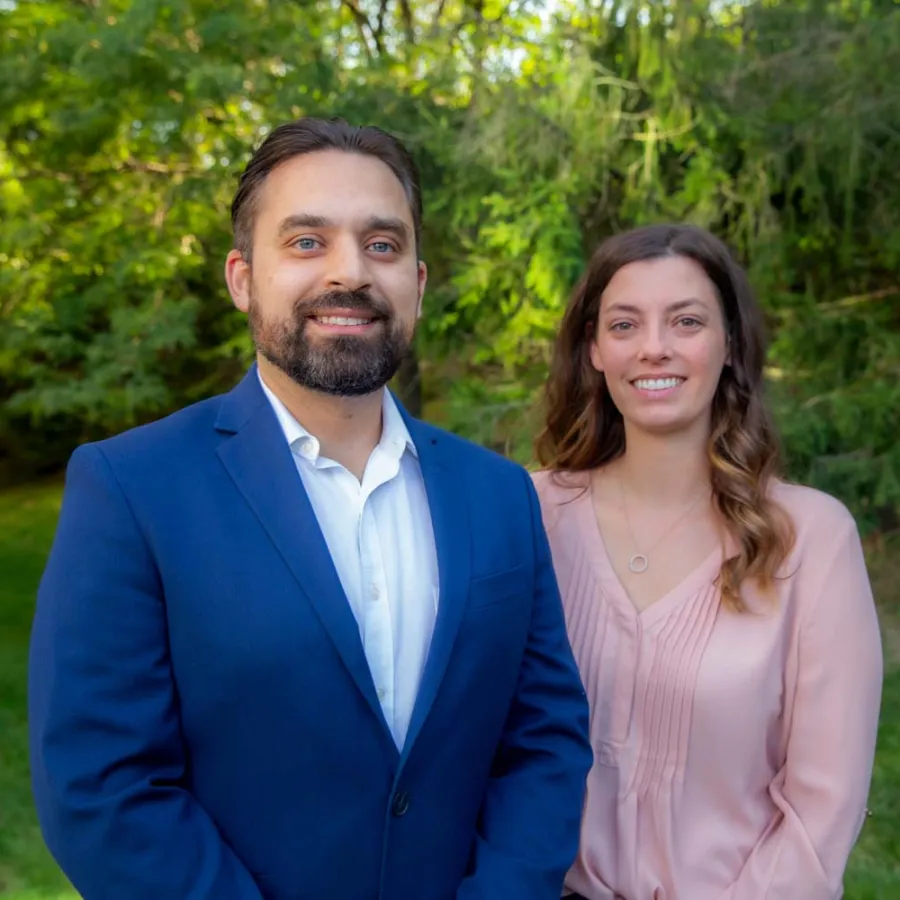 Smiling man in blue suit and woman in pink blouse standing outdoors with green trees and grass in background