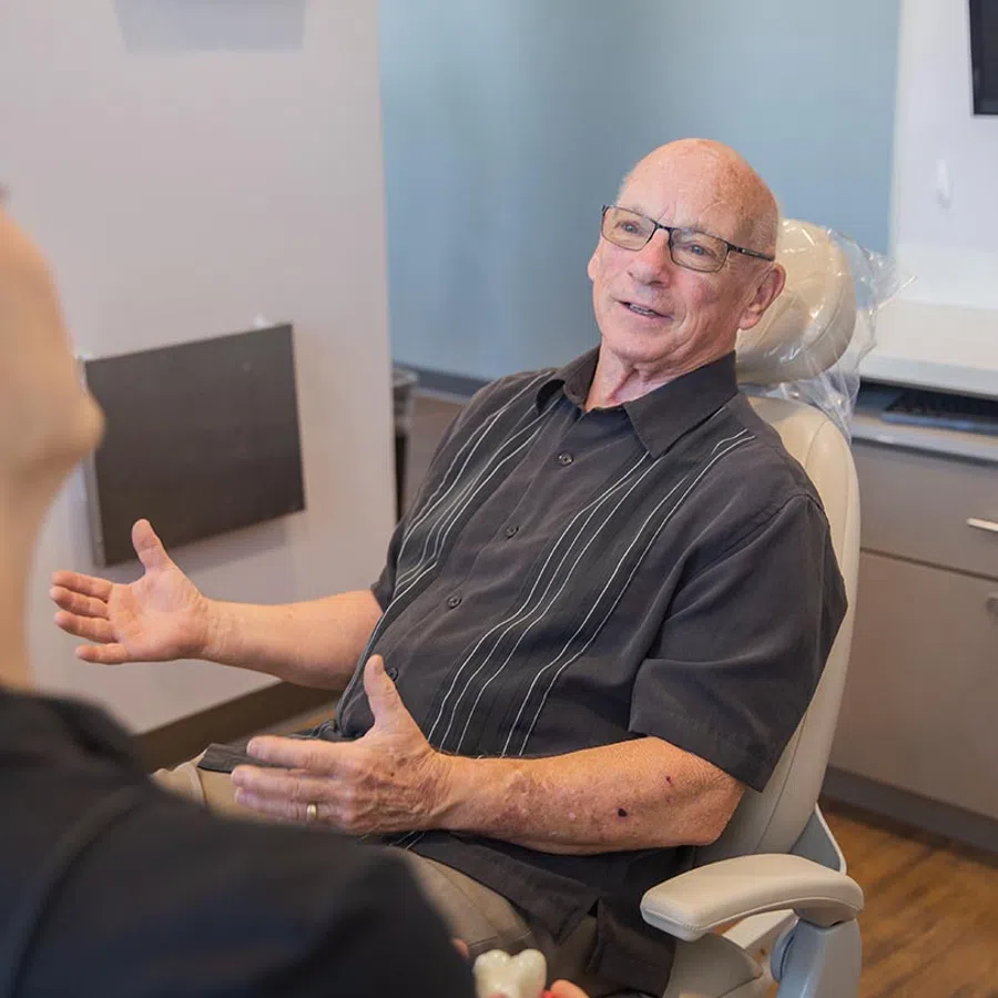 Elderly man with glasses sitting in a chair and talking to another person in a room with light walls.
