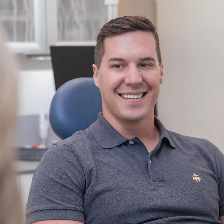 Young man in gray polo shirt smiling while sitting in a dental office chair during a consultation.
