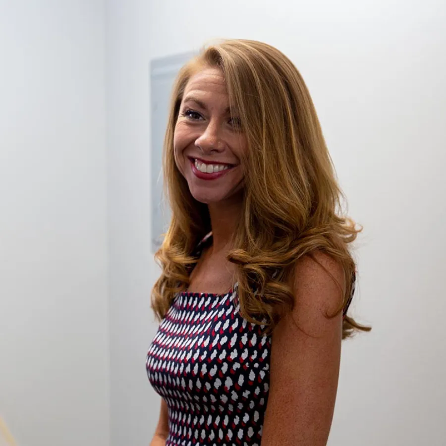 Smiling woman with long curly blonde hair wearing a patterned sleeveless dress against a plain light background