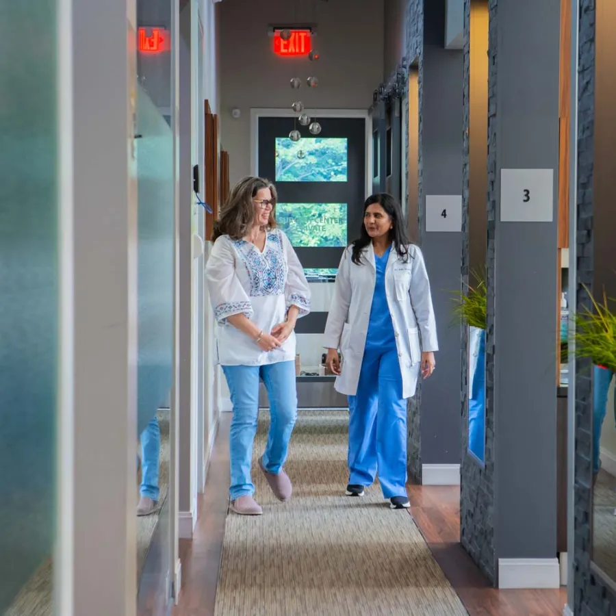 Female doctor in white coat walking and talking with female patient in a modern medical clinic hallway.