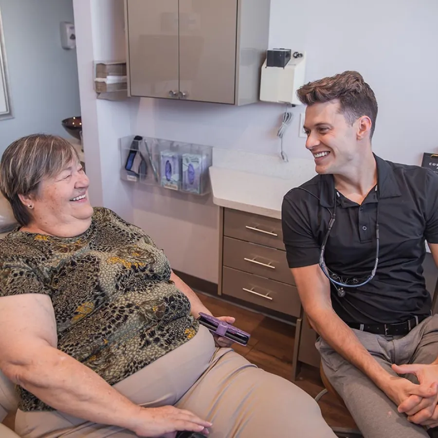 Smiling female dentist in black scrubs sitting at desk in a modern dental office with dental chair and equipment.