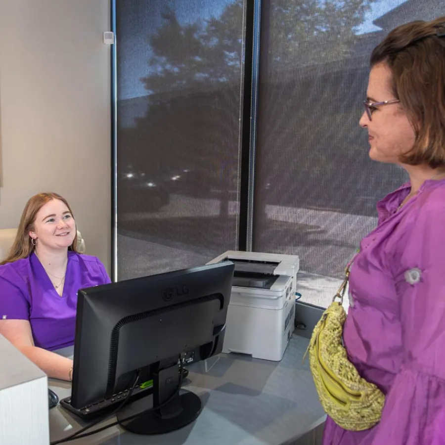 Smiling receptionist in purple scrubs assists woman in purple dress with yellow bag at front desk in office.