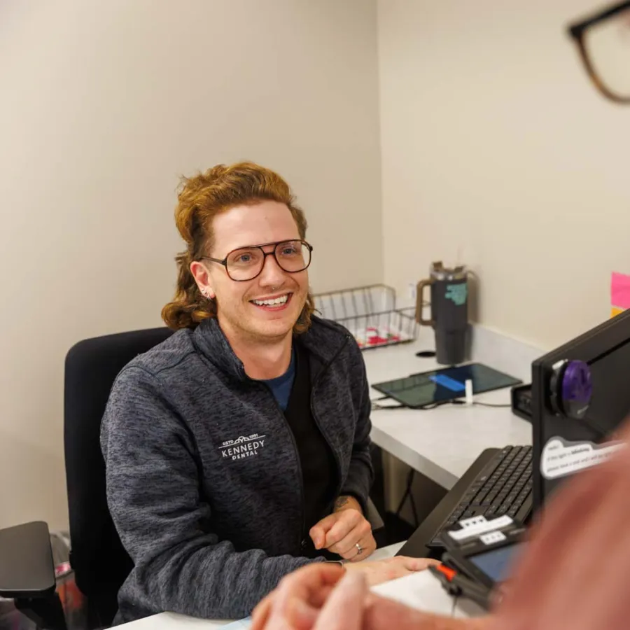 Smiling woman in glasses and jacket assisting an older man at an office desk with computer and paperwork.