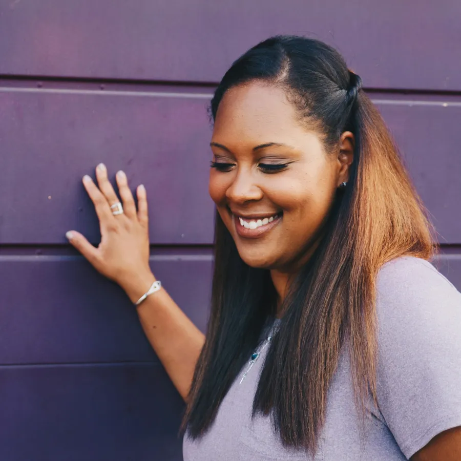 Smiling woman with long hair resting hand on purple wall, wearing casual light purple top and silver jewelry.