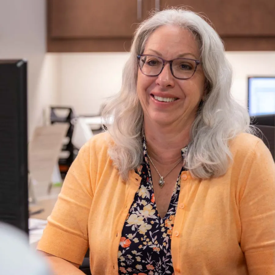 Smiling woman in glasses and orange cardigan talking to a man in an office setting.