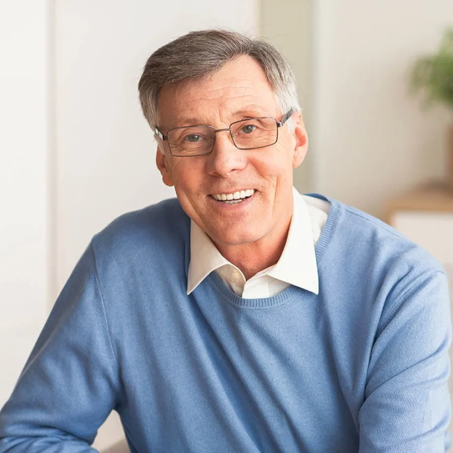 Smiling middle-aged man with glasses wearing a blue sweater and white shirt sitting indoors in a bright room.