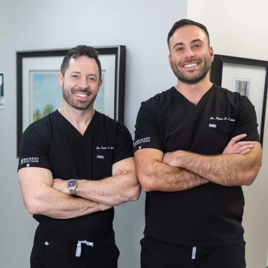 Two male doctors in black scrubs posing with arms crossed, smiling in a medical office setting.