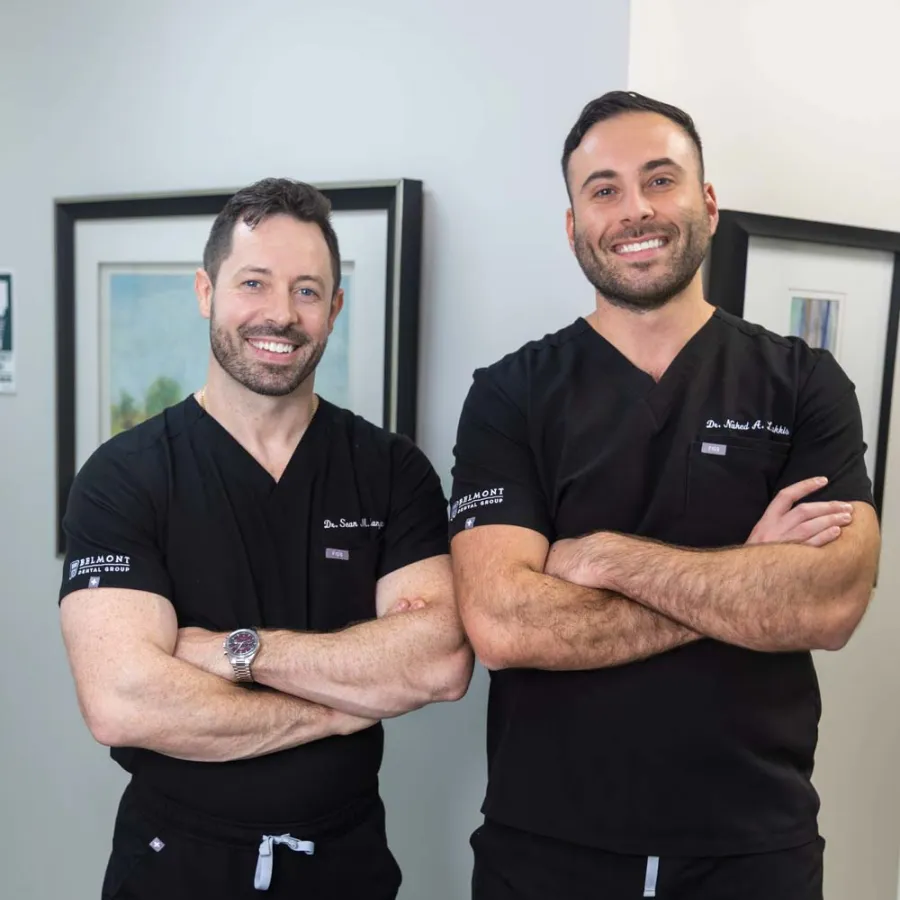 Two male doctors in black scrubs posing with arms crossed, smiling in a medical office setting.