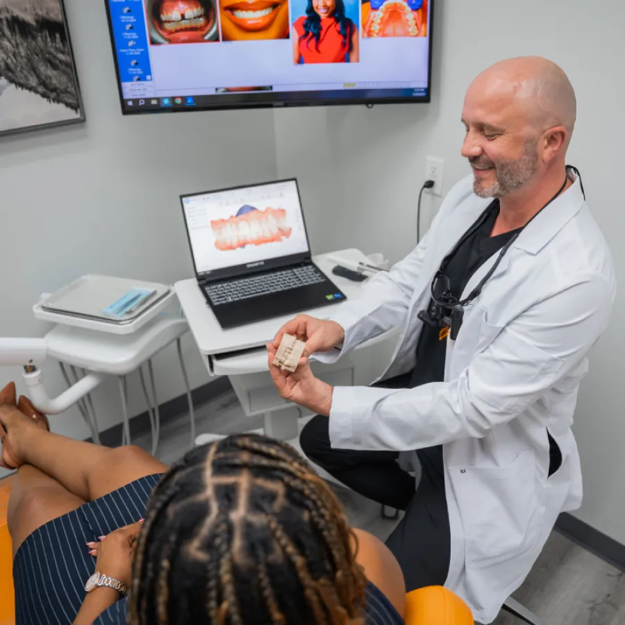 Dentist in white coat showing dental mold to patient in modern dental office with laptop and screen.