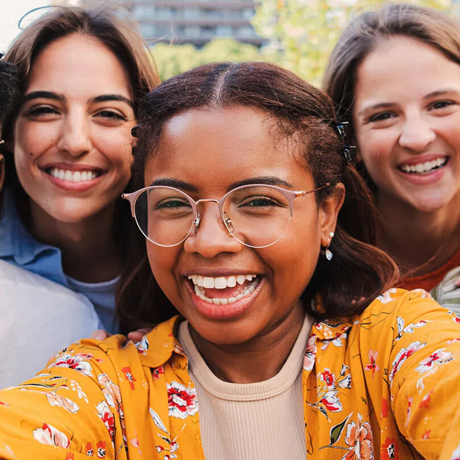 Three diverse young women smiling happily outdoors taking a close-up selfie together.
