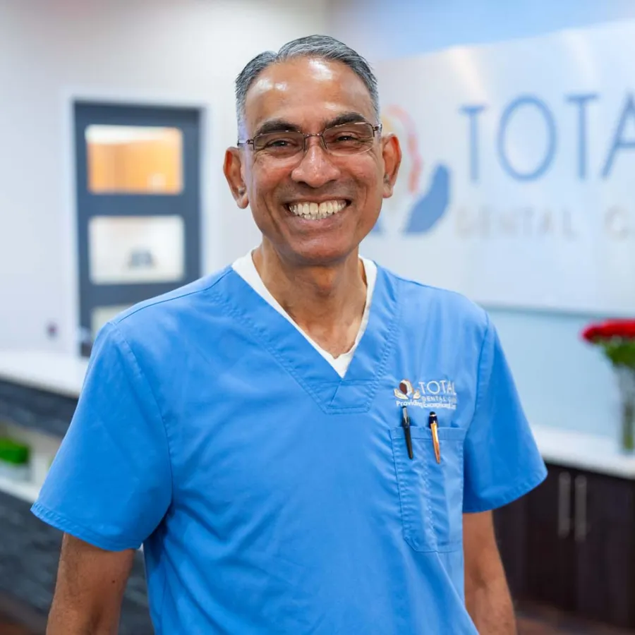 Smiling male dentist in blue scrubs standing in Total Dental Care clinic with a bright, welcoming atmosphere.