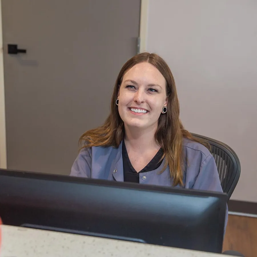 Smiling woman with brown hair in casual uniform sitting behind a desk with a computer monitor.