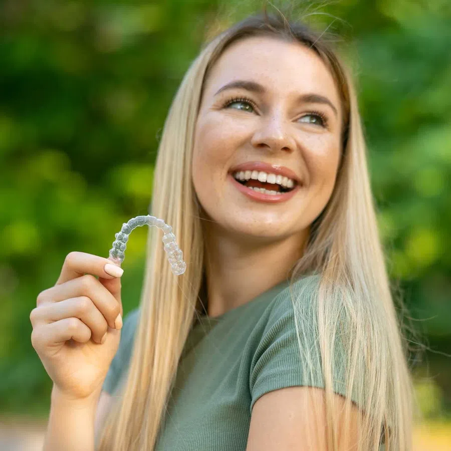 Smiling young woman holding a clear dental aligner outdoors with green blurred background