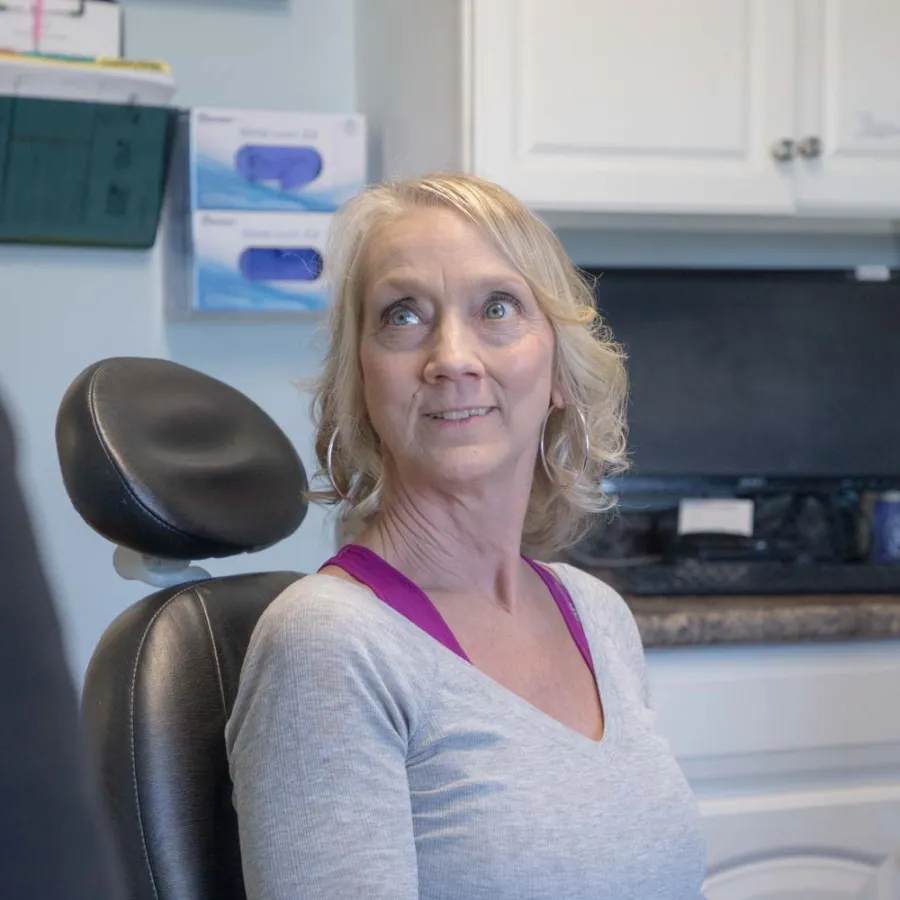 Middle-aged woman sitting in a dental chair looking toward dentist in a modern clinic room.