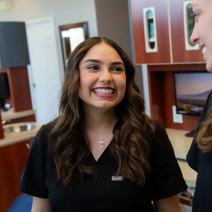 Two female healthcare professionals smiling and talking in a medical office with computer monitors and cabinets.