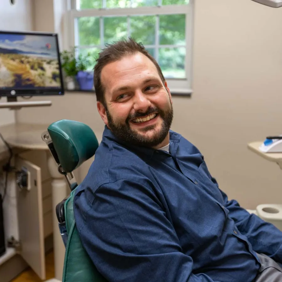 Smiling man sitting in a dental chair in a bright dental office with equipment and monitor visible.