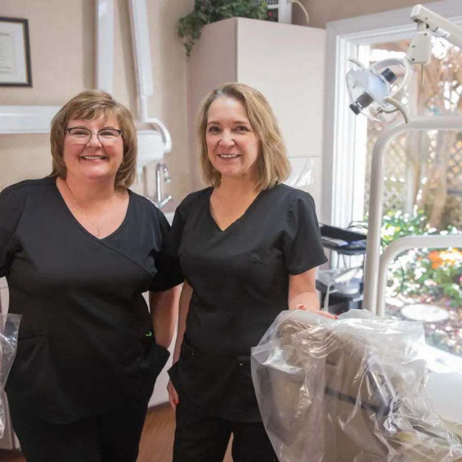 Two female dental hygienists in black scrubs smiling inside a bright dental office with equipment and window view.