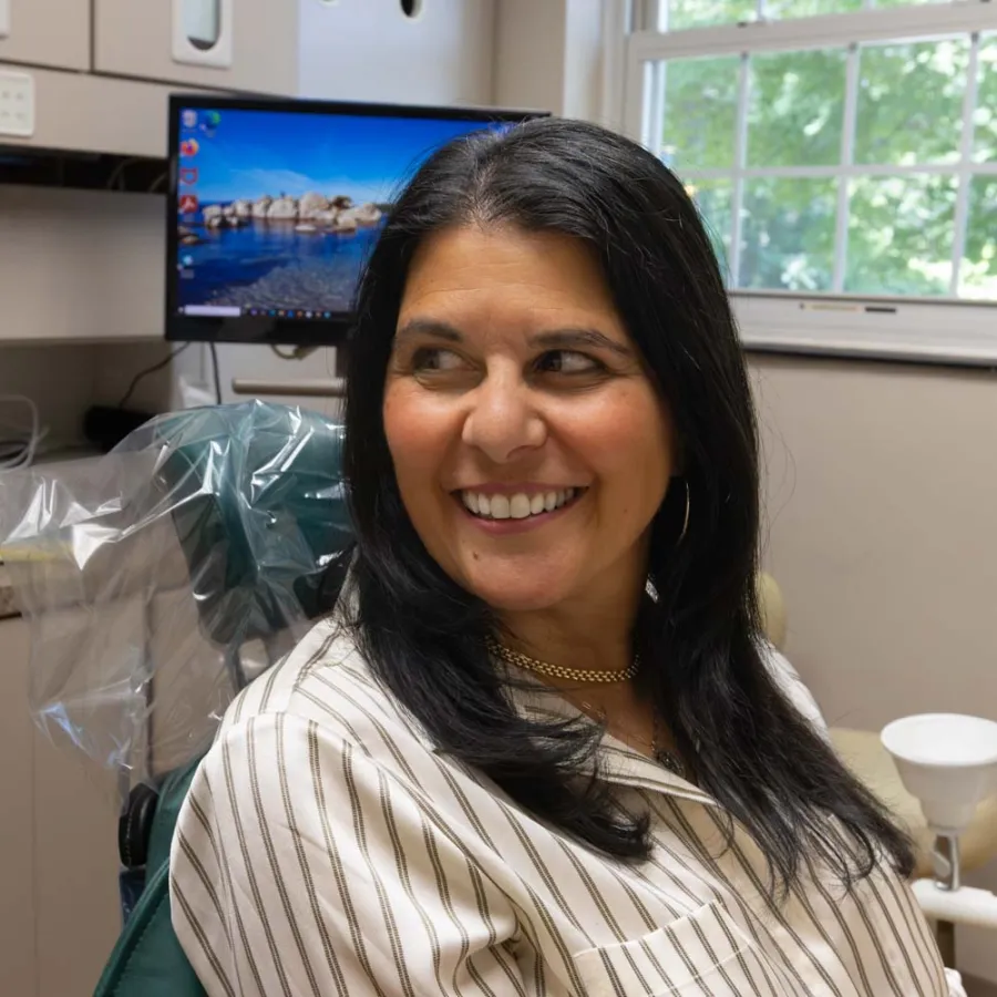Smiling woman sitting in a dental chair in a bright dental office with a computer screen and window in background