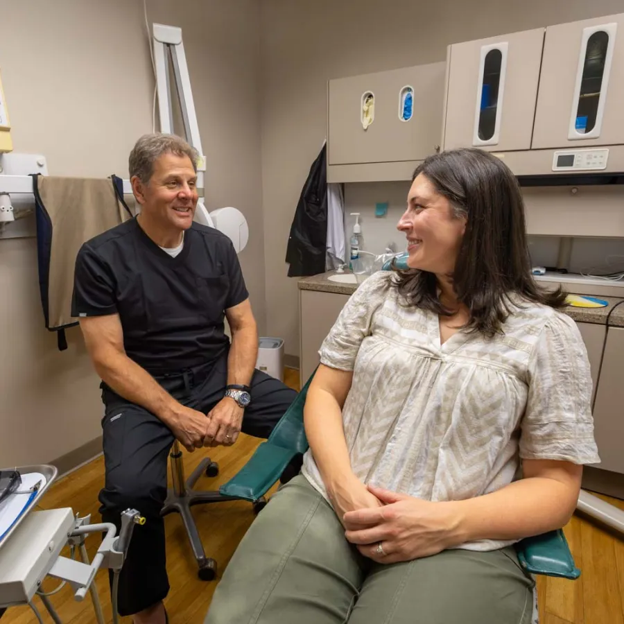 Dentist in black scrubs talking and smiling with a female patient in dental chair inside a clinic room.