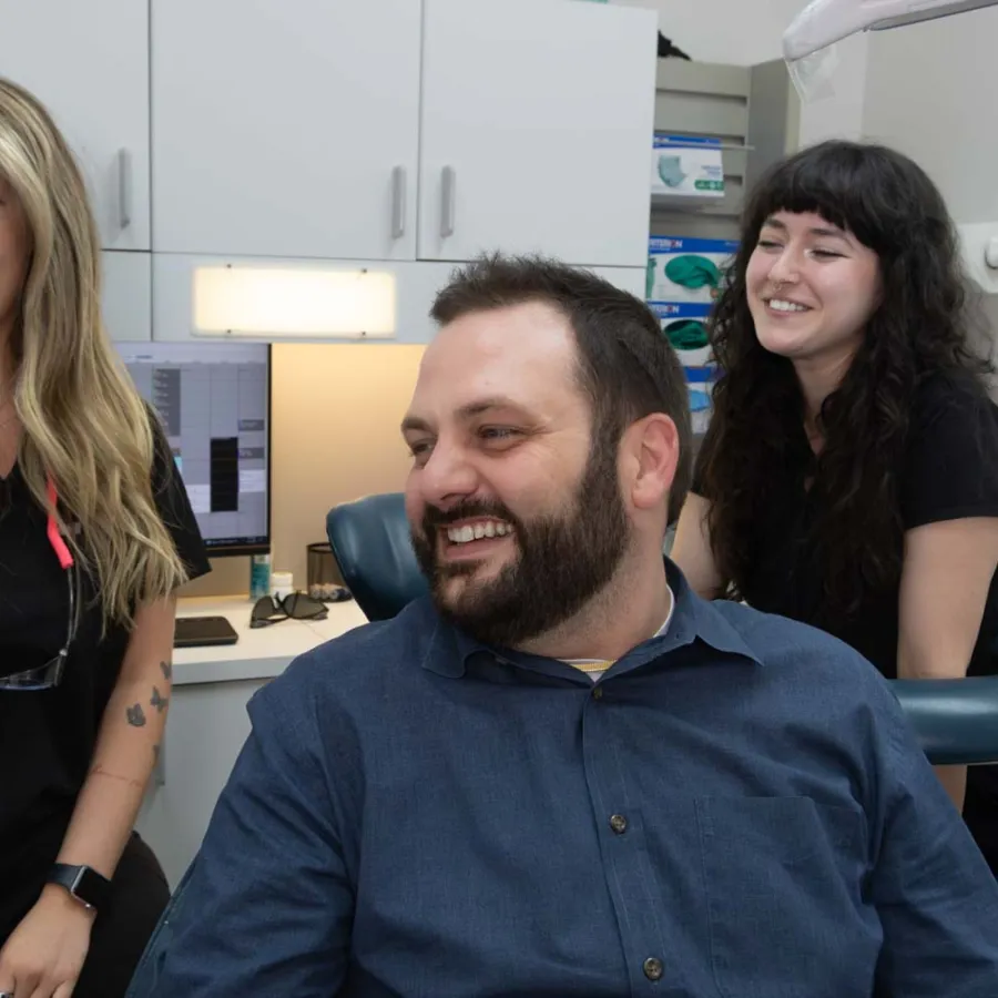 Smiling male patient and two female dental staff in a modern dental office with computer and medical supplies.