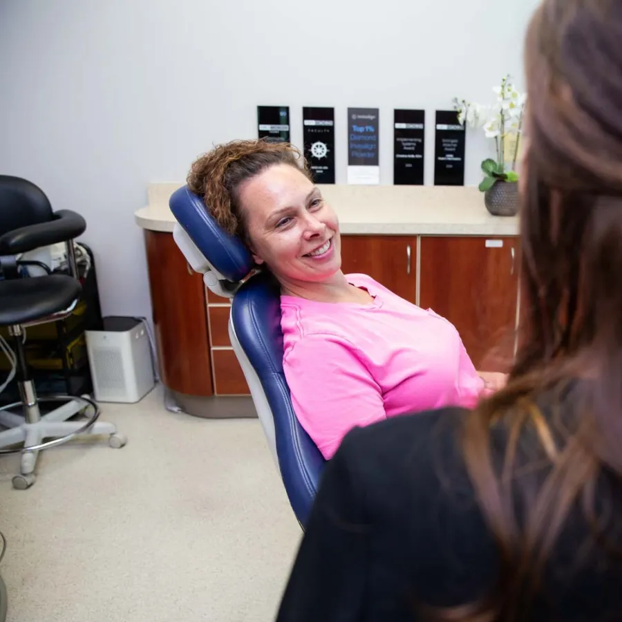 Woman in pink shirt smiling while sitting in a dental chair during a consultation with a dental professional.