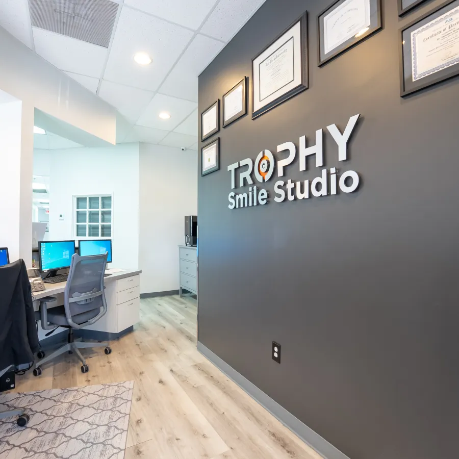 Modern dental office interior with Trophy Smile Studio logo, computer desks, chairs, and certificates on wall.