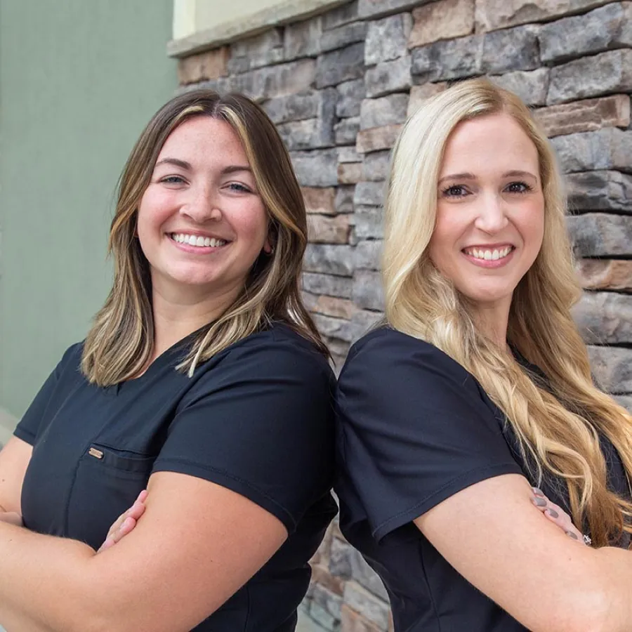 Two smiling women in black scrubs stand back-to-back with arms crossed in front of a stone wall.