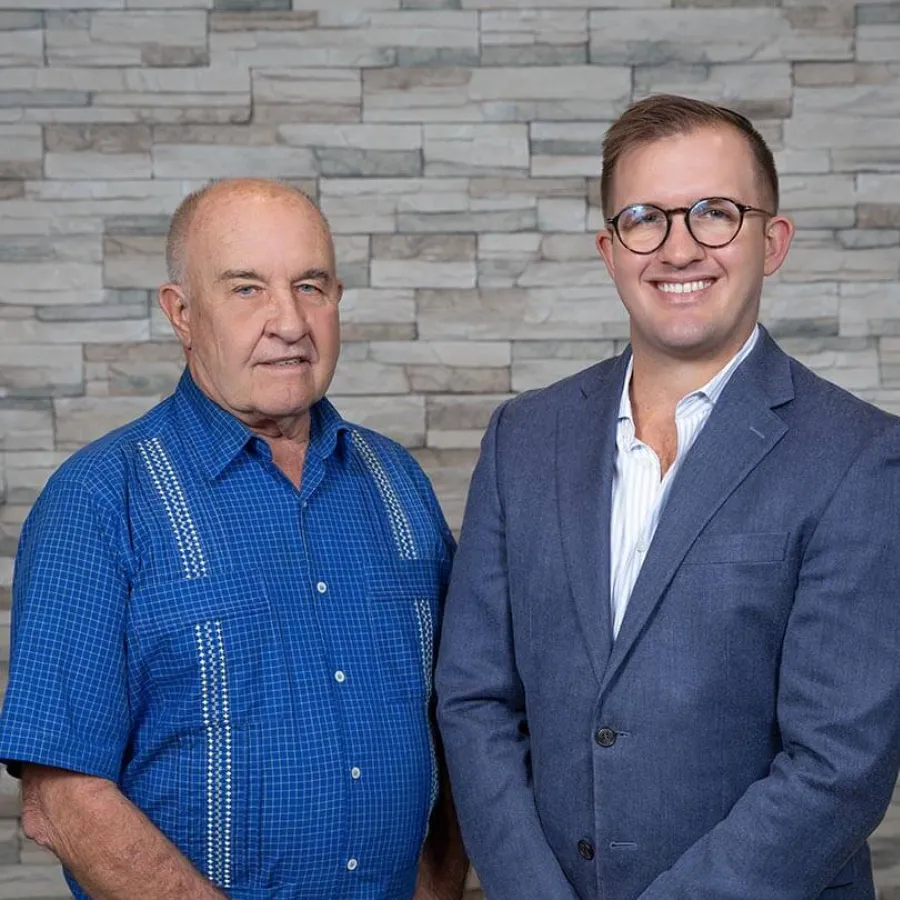 Older man in blue shirt and younger man in suit stand smiling against stone wall background.